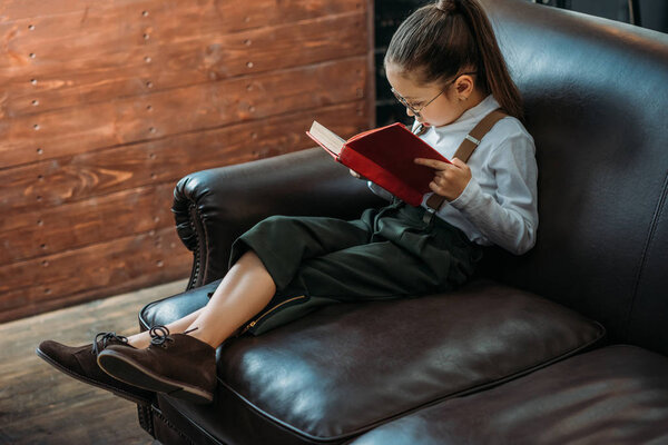 stylish little child reading book on couch at home