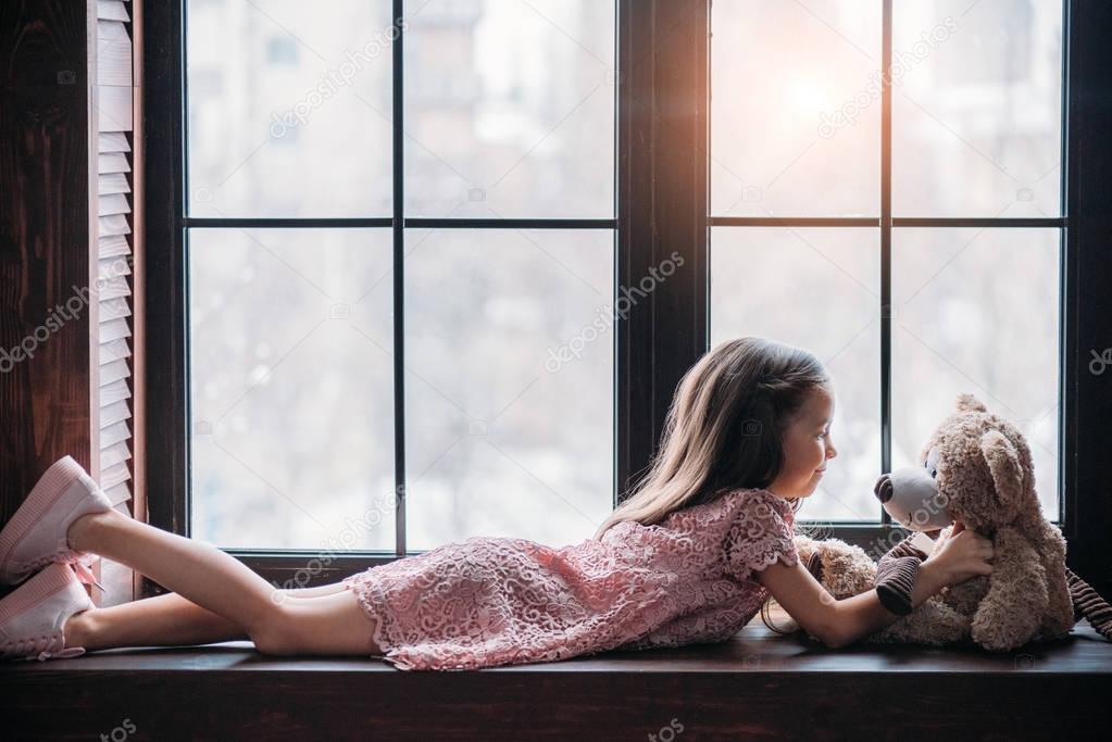 side view of little child playing with teddy bear while lying on windowsill