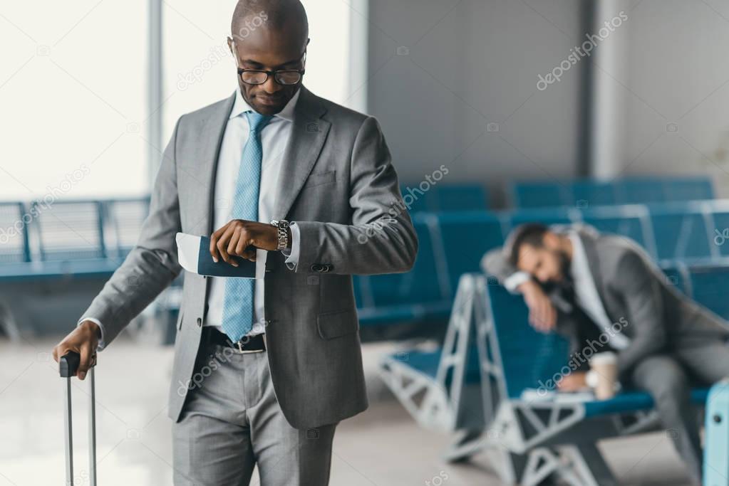 handsome businessman looking at watch while waiting for flight in airport lobby with sleeping man blurred on background