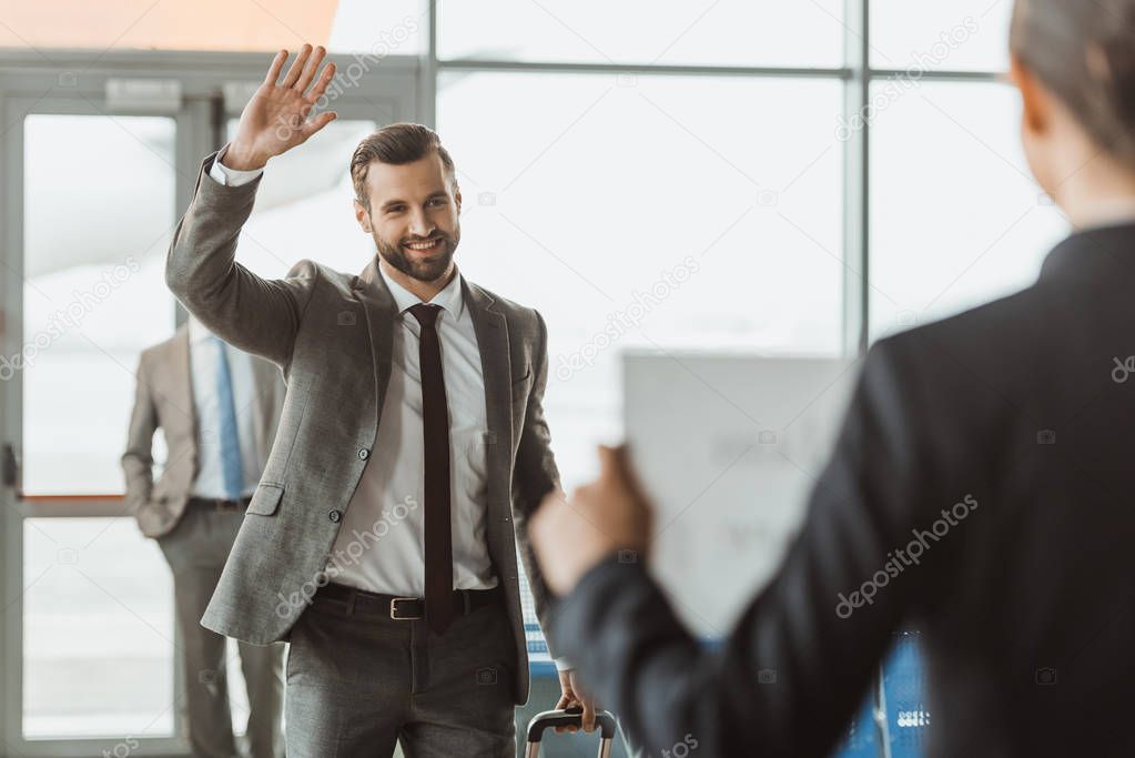 businessman waving to partner who waiting for him with name sign on paper at airport