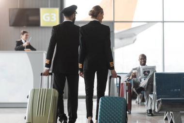 back view of male and female pilots walking by airport lobby with suitcases clipart