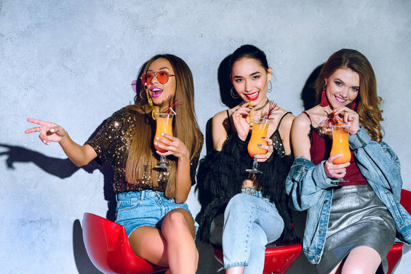 счастливые стильные мультиэтнические девушки сидят на барных стульях и пьют коктейли на вечеринке
