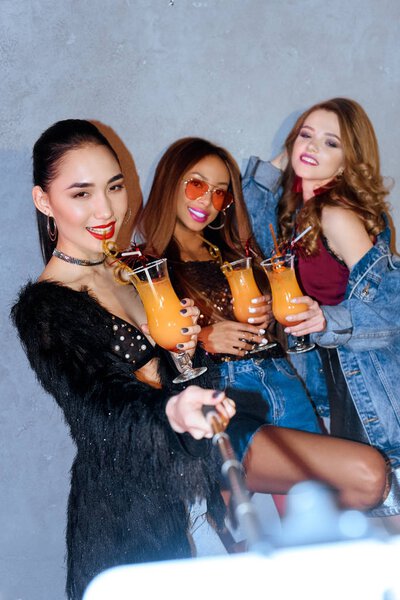 stylish multiethnc girls drinking cocktails and taking selfie with smartphone at party