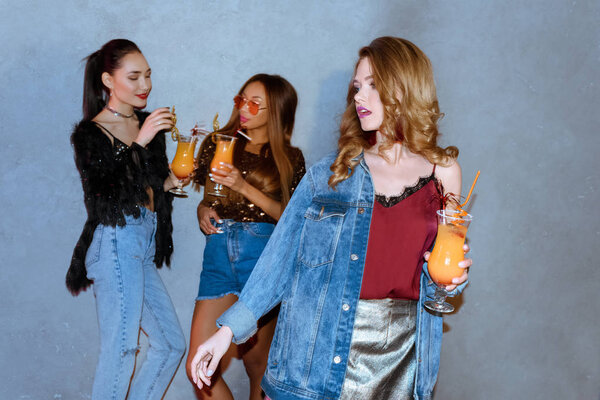 fashionable girl holding glass with cocktail while multiethnic friends standing behind