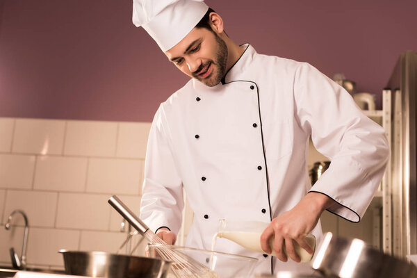 portrait of confectioner in chef hat pouring milk into bowl while making dough in restaurant kitchen