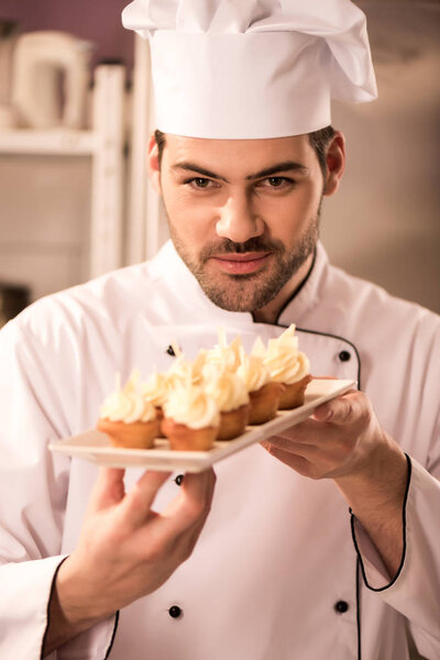 portrait of young confectioner looking at cupcakes on plate in hands