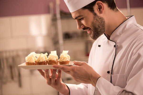 side view of smiling confectioner looking at cupcakes on plate in hands