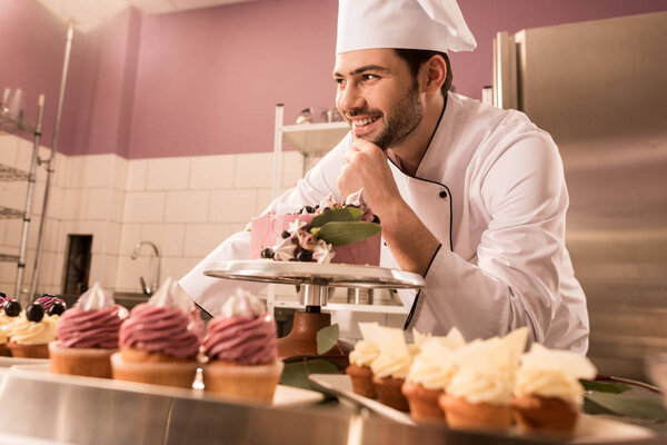 smiling confectioner standing at counter with cake and cupcakes in restaurant kitchen