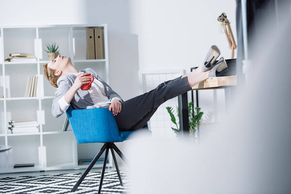 fashionable young businesswoman relaxing with cup of coffee in chair at office