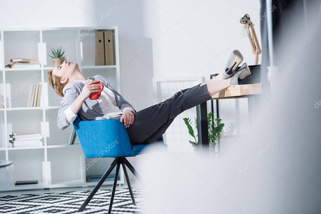 fashionable young businesswoman relaxing with cup of coffee in chair at office