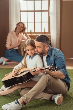 Baba ve kızı içinde oturan annesi ile karton evin önünde çimenlerin üzerinde gitar çalmak