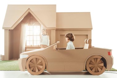 kid riding cardboard car in front of house while girlfriend waving to him isolated on white clipart