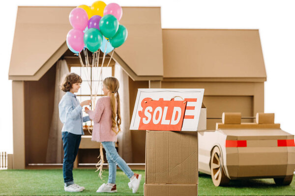 kid presenting balloons to girlfriend in front of cardboard house with sold signboard on foreground isolated on white
