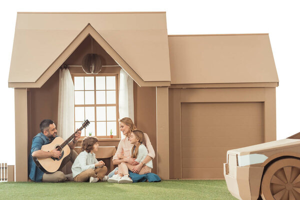 father playing guitar for kids and wife at new cardboard house isolated on white