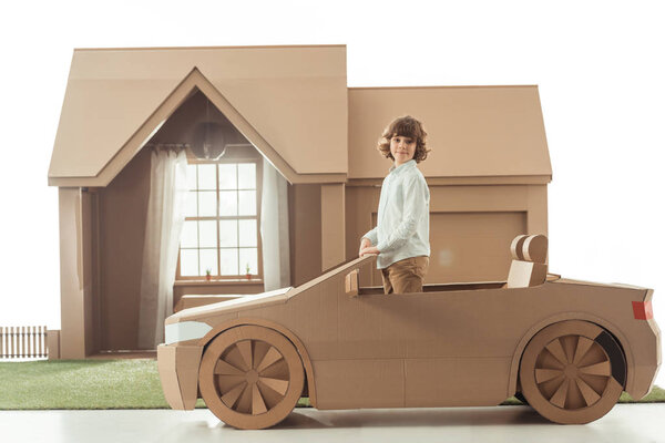 little kid standing in cardboard car in front of cardboard house isolated on white