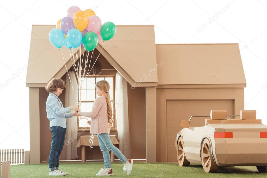 kid presenting balloons to girlfriend in front of cardboard house isolated on white