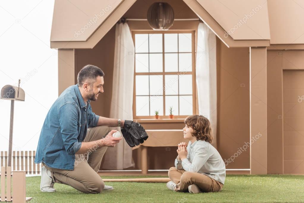father teaching his son how to play baseball on grass in front of cardboard house