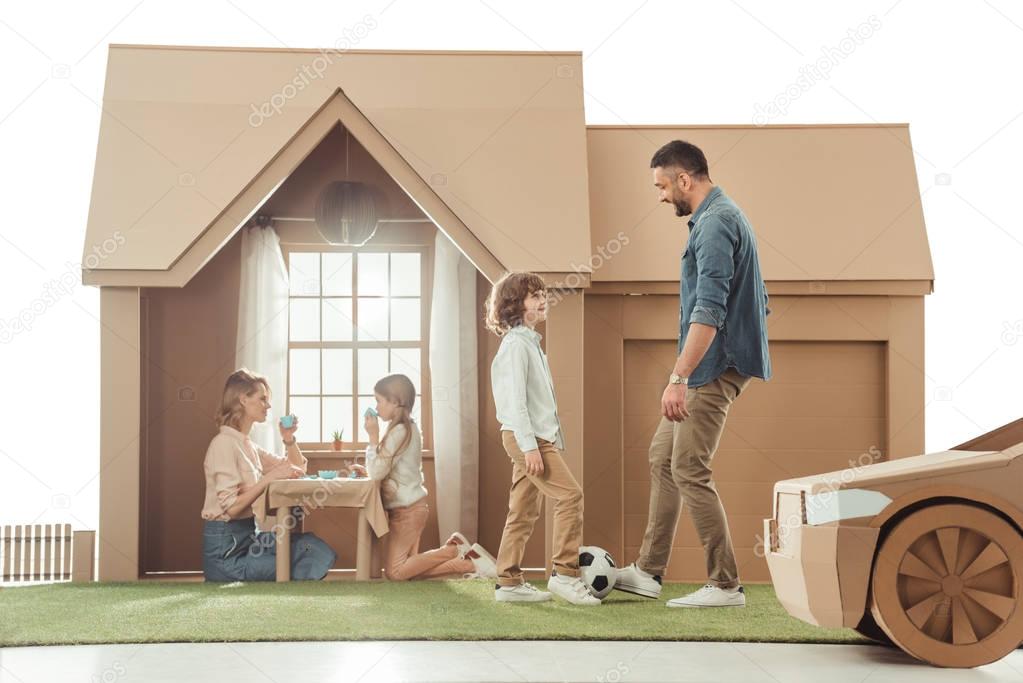 young father teaching his som to play soccer on yard of cardboard house isolated on white