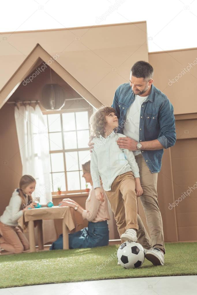 father teaching his som to play soccer on yard of cardboard house