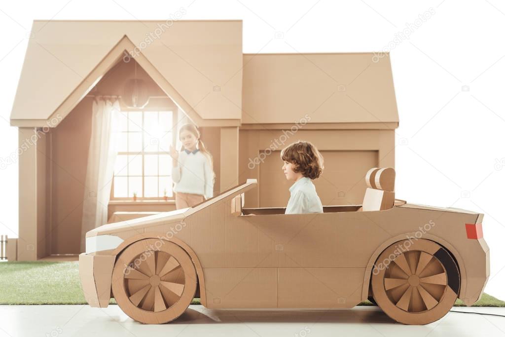 kid riding cardboard car in front of house while girlfriend waving to him isolated on white