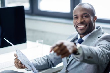 smiling african american businessman with newspaper pointing away at workplace