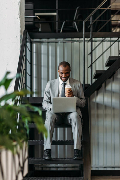 african american businessman with coffee to go using laptop while sitting on stairs 