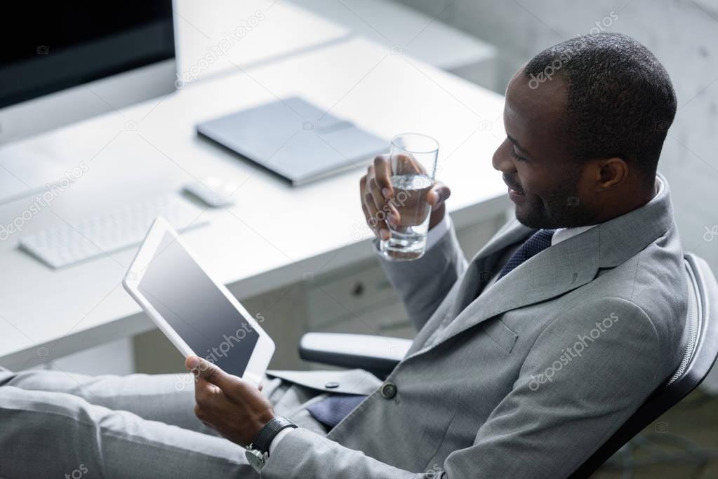 african american businessman with glass of water using tablet at workplace in office