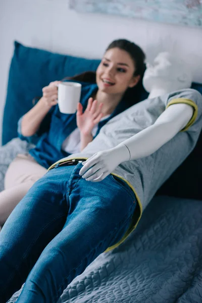 young woman with cup of coffee talking to mannequin while resting on bed, unrequited love concept