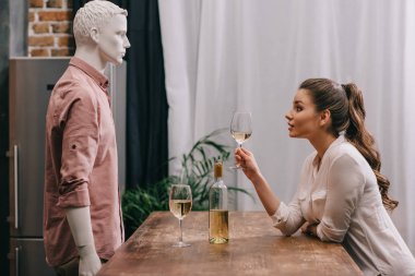 side view of young woman with glass of wine at table with male manikin, unrequited love concept clipart