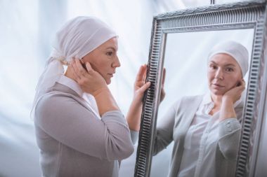 serious sick mature woman in kerchief looking at mirror, cancer concept clipart