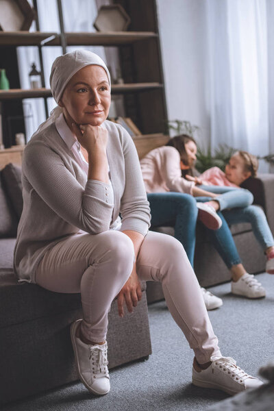 pensive sick mature woman in kerchief looking away while family members having fun behind 