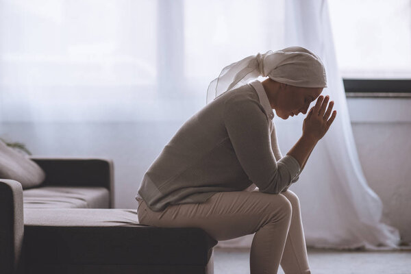 side view of upset sick mature woman in kerchief sitting at home, cancer concept