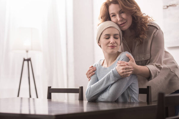 mature woman hugging and supporting sick smiling daughter in kerchief