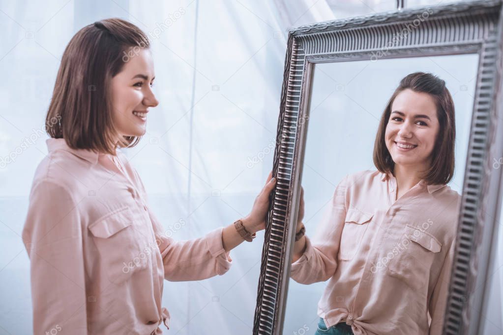 beautiful smiling young woman standing near mirror and looking at reflection