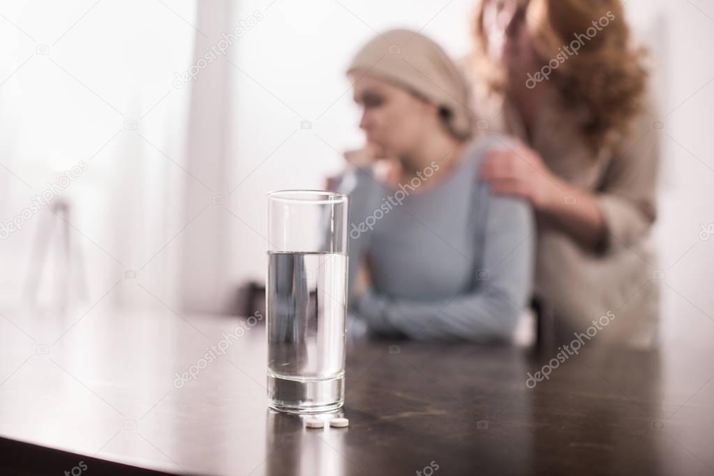 close-up view of glass of water and woman supporting sick daughter in kerchief behind