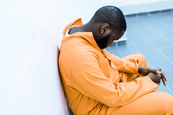 sad african american prisoner sitting on bench in prison cell