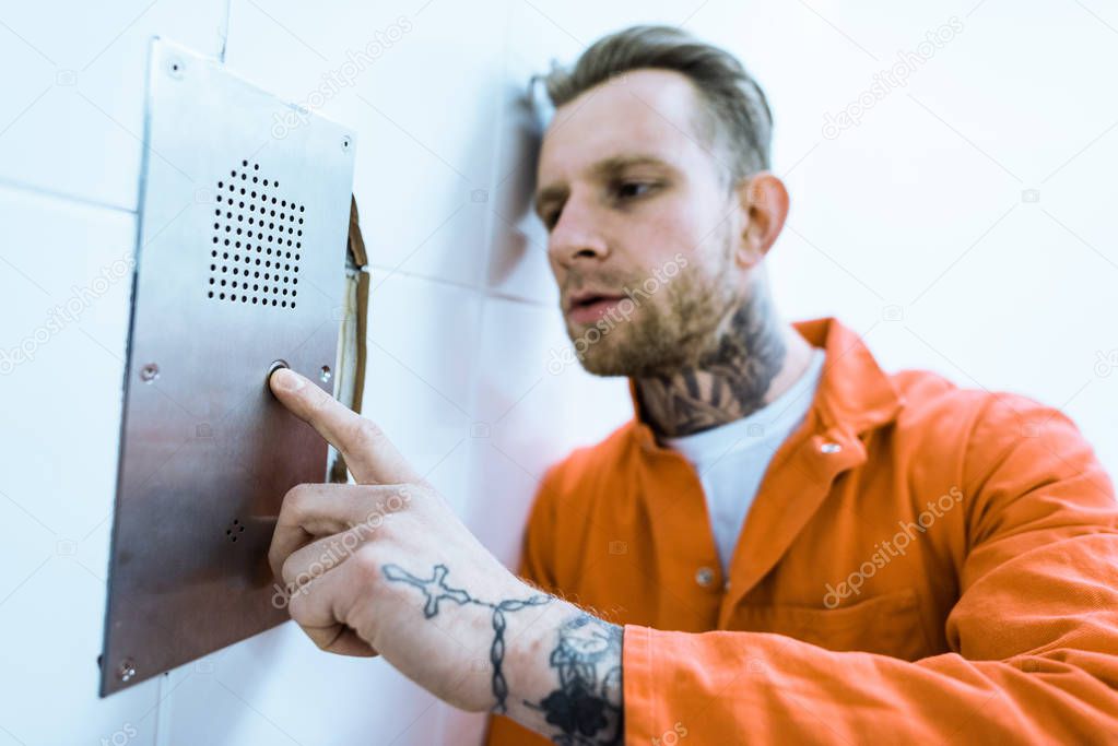 tattooed criminal in orange uniform pressing button in prison cell