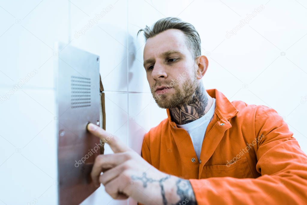 tattooed prisoner in orange uniform pressing button in prison cell