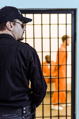 rear view of prison officer standing near prison cell with multicultural inmates clipart