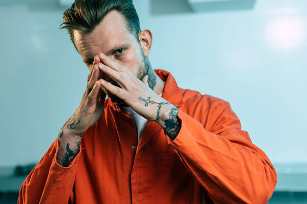 prisoner with tattoos covering face with hands and looking at camera