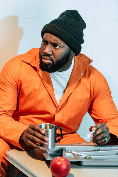 african american prisoner holding cup and fork in prison cell
