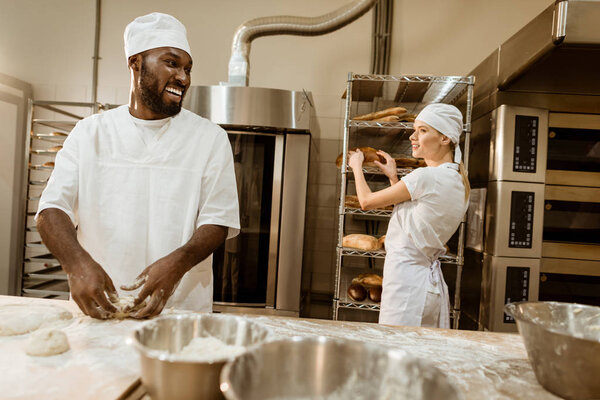 african american baker kneading dough at baking manufacture and chatting with colleague