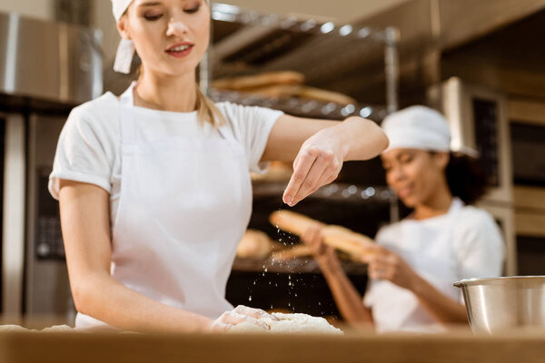 happy female baker kneading dough and pouring flour on it at baking manufacture while her colleague working blurred on background