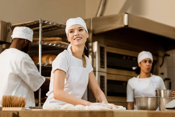 若い女性ベイカー背景にぼやけながら作業している彼女の同僚の製造をパンで生地を練り — ストック写真
