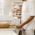 Красивый пекарь, работающий с промышленным тестовым роликом при производстве выпечки Красивый пекарь, работающий с промышленным тестовым роликом при производстве выпечки