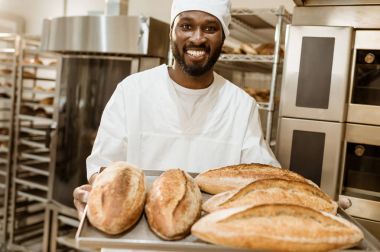 handsome african american baker with tray of fresh loaves of bread on baking manufacture clipart