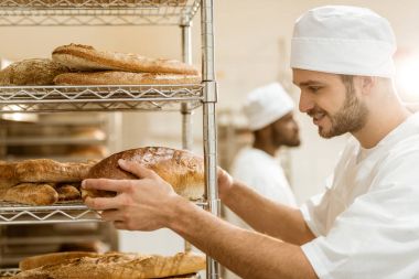 handsome smiling  baker putting fresh bread loaf on shelf at baking manufacture clipart