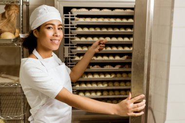 smiling female baker pointing at dough inside of industrial oven at baking manufacture clipart