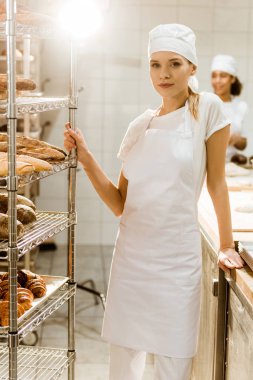 young female baker standing near shelves with fresh loaves of bread at baking manufacture clipart