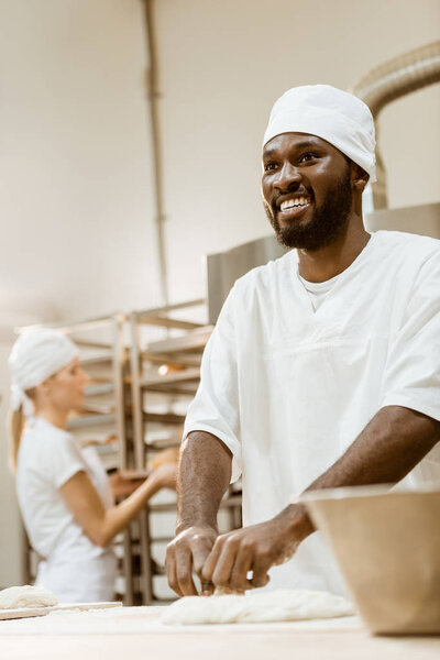 happy african american baker kneading dough while his female colleague working on background at baking manufacture